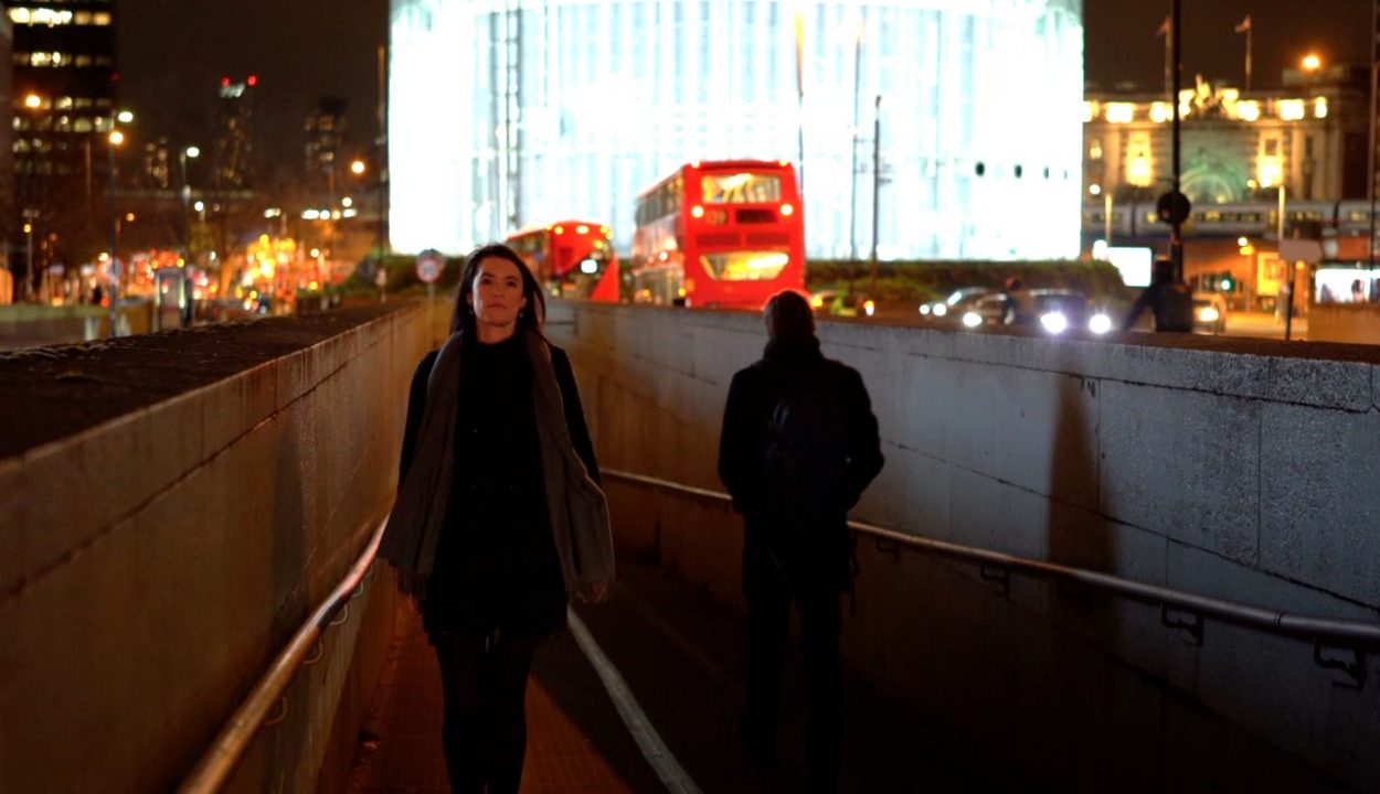 A woman walks through a dimly lit underpass at night, with a red double-decker bus and a brightly illuminated circular building in the background. The city lights add a vibrant glow to the scene.