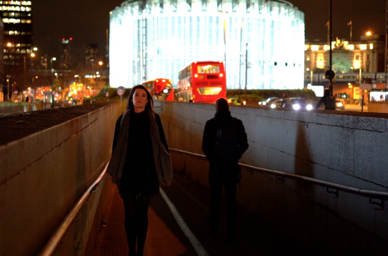 A woman walks through a dimly lit underpass at night, with a red double-decker bus and a brightly illuminated circular building in the background. The city lights add a vibrant glow to the scene.