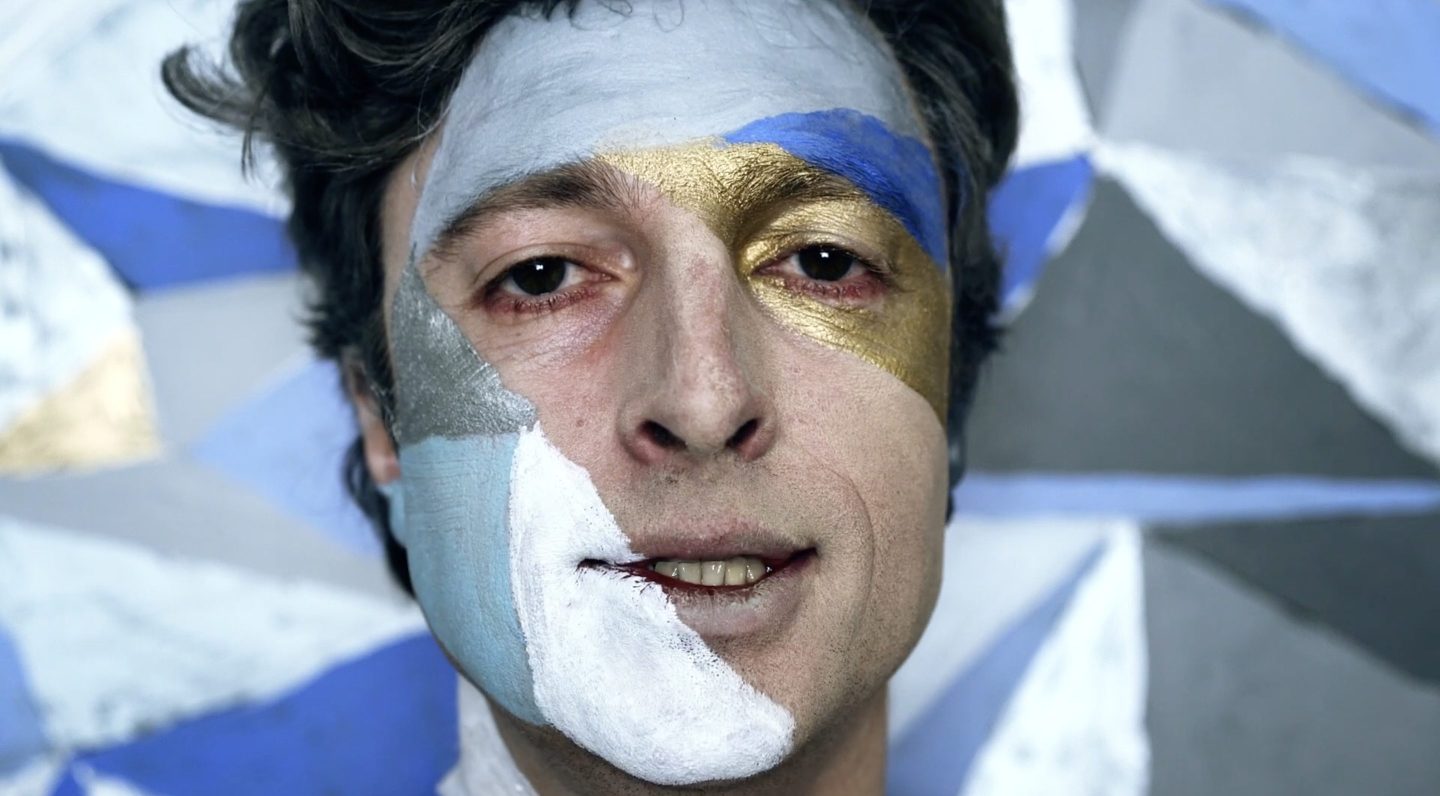 A person with face paint in blue, white, and gold is smiling slightly. The background features geometric shapes in matching colours.