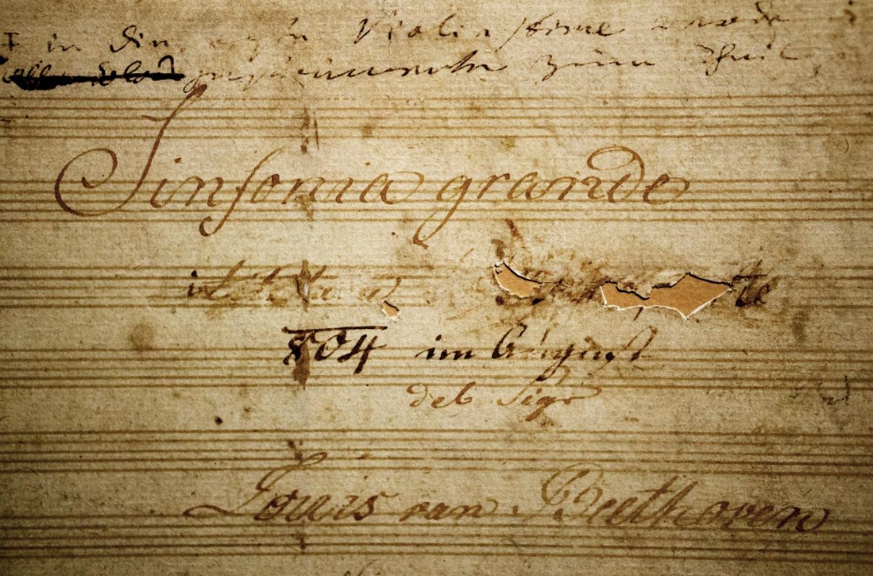 A close-up of a music manuscript titled "Sinfonia Grande." The aged paper shows handwritten notes and text, including the name "Ludwig van Beethoven." There are visible creases and a torn section on the document.