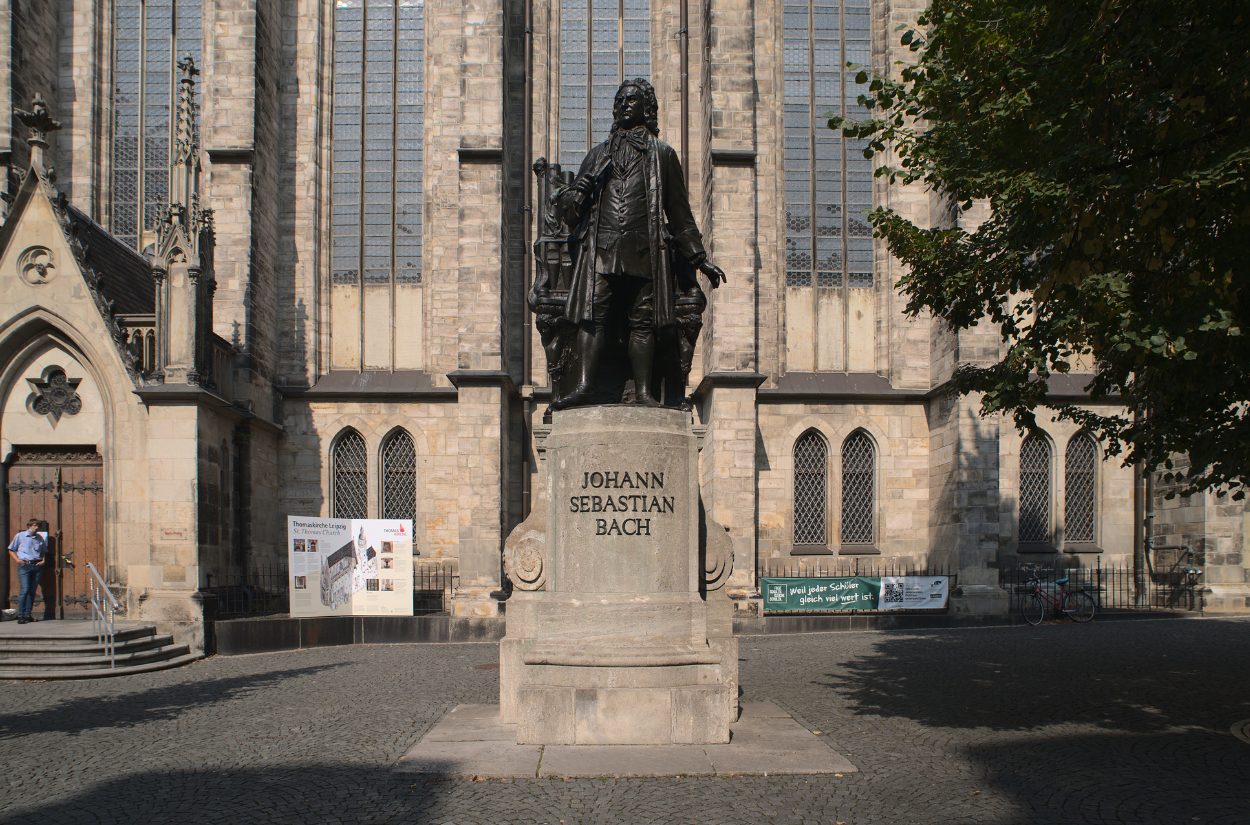 A statue of Johann Sebastian Bach stands in front of a historic stone building with tall windows. The base of the statue displays his name. A tree is on the right, and a few people are in the background.