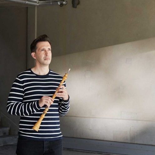 Daniel Bates stands to the left of the image, looking towards the right. He wears a black and white striped shirt and holds an oboe in both hands. There is a plain wall behind him, and light filters in from the right.