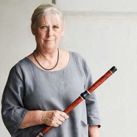 Lisa Beznosiuk wears a grey long sleeved jumper and holds a flute in her left hand. Behind her is a plain white wall.