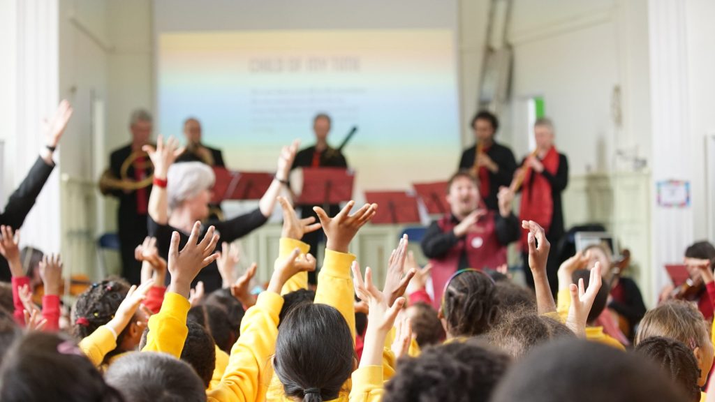 A group of children in yellow shirts raise their hands enthusiastically during a music performance. In the background, several musicians play instruments and a screen displays information about the song. A conductor directs the children from the front.