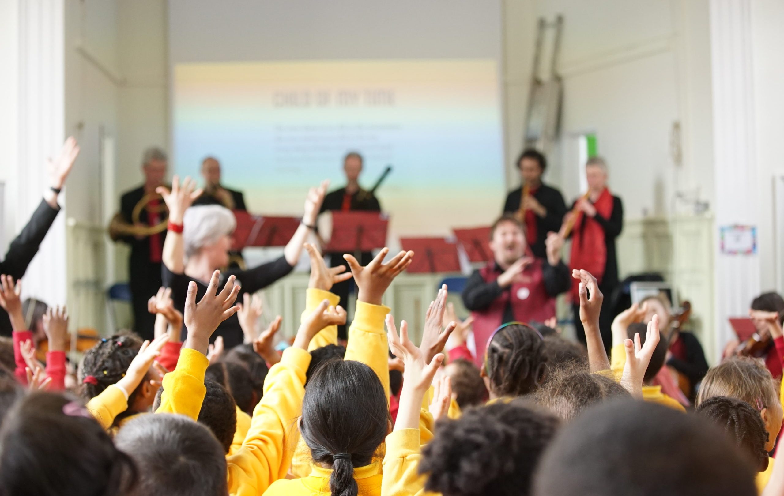 A group of children in yellow shirts raise their hands enthusiastically during a music performance. In the background, several musicians play instruments and a screen displays information about the song. A conductor directs the children from the front.