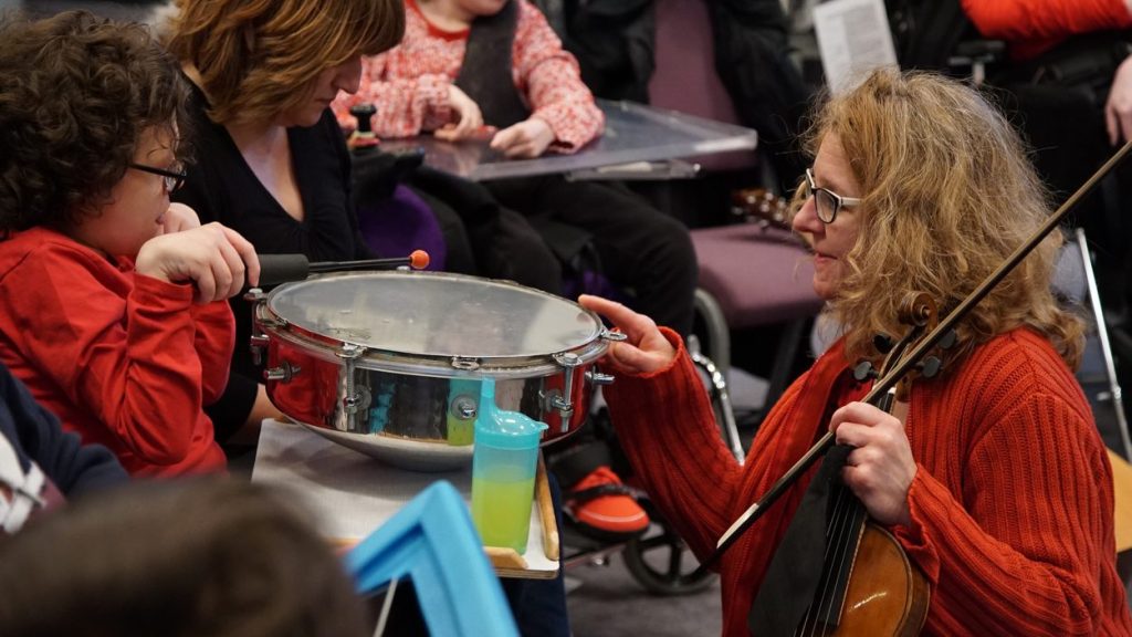 A woman in a red sweater holds a violin and interacts with a child playing a snare drum. The child, also wearing a red outfit, uses a drumstick to tap the drum. Other children and caregivers are visible in the background.