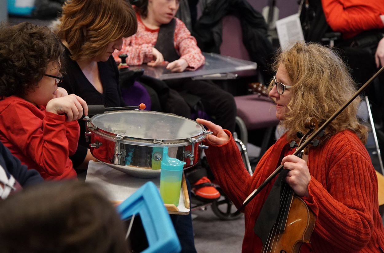 A woman in a red sweater holds a violin and interacts with a child playing a snare drum. The child, also wearing a red outfit, uses a drumstick to tap the drum. Other children and caregivers are visible in the background.