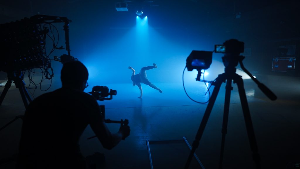 A student dancer performs a break-dancing move under dramatic blue lighting while being filmed by a cameraman. Silhouetted camera equipment stands to the side, recording the action unfold on a dimly lit stage.