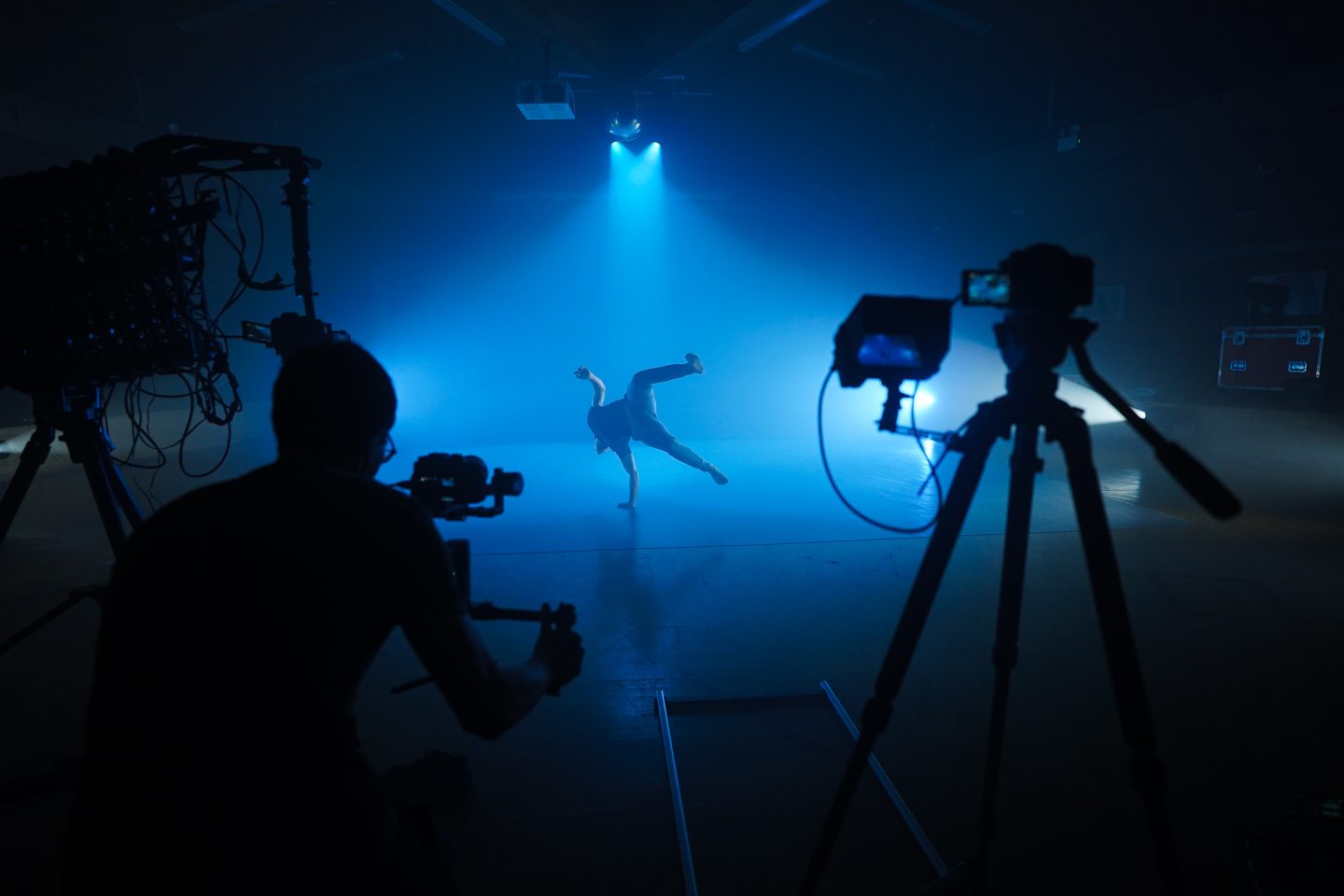 A student dancer performs a break-dancing move under dramatic blue lighting while being filmed by a cameraman. Silhouetted camera equipment stands to the side, recording the action unfold on a dimly lit stage.