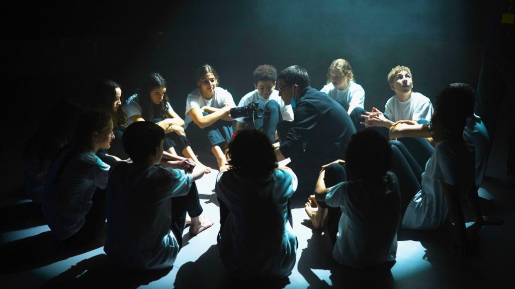 A group of people seated in a circle illuminated by a spotlight in an otherwise dark space. The individuals, dressed in casual clothes, seem engaged in an intense discussion or storytelling session. Some people are sitting cross-legged, while others are in various relaxed postures.