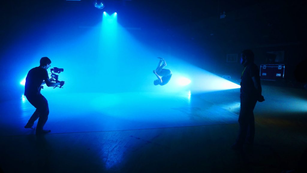 A student dancer performs an acrobatic flip under dramatic blue lighting while being filmed by a cameraman. Silhouetted onlookers stand to the side, watching the action unfold on a dimly lit stage.