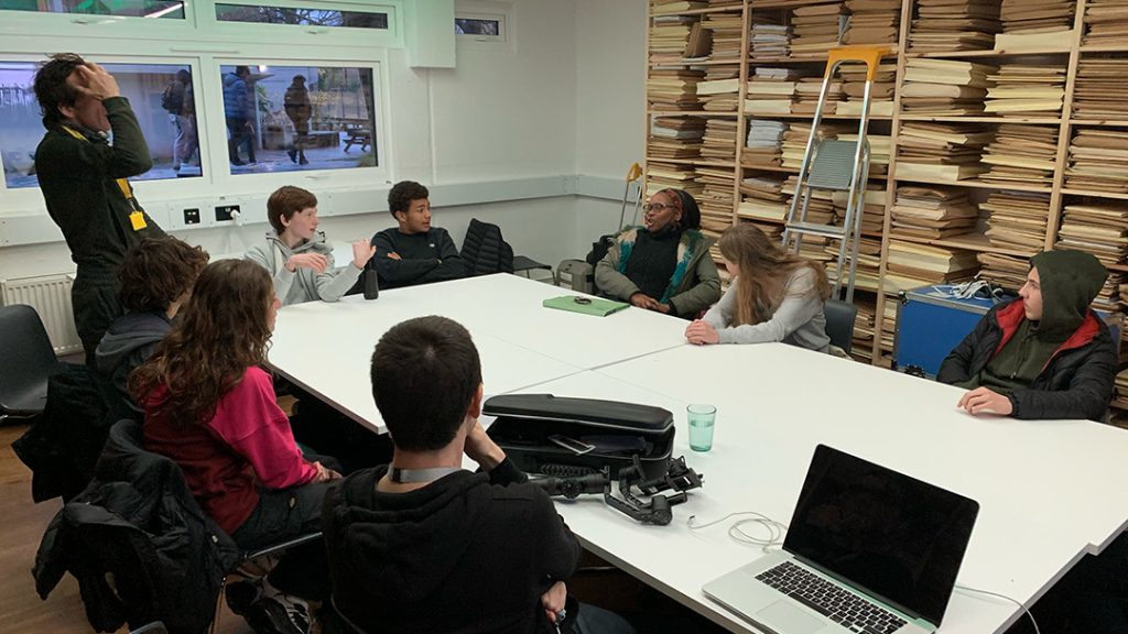 A group of people sits around a large white table in a brightly lit room. The table has a laptop and a camera on it. One person is standing, while others are seated, engaged in conversation. Stacks of papers or files are organised on shelves against the wall.