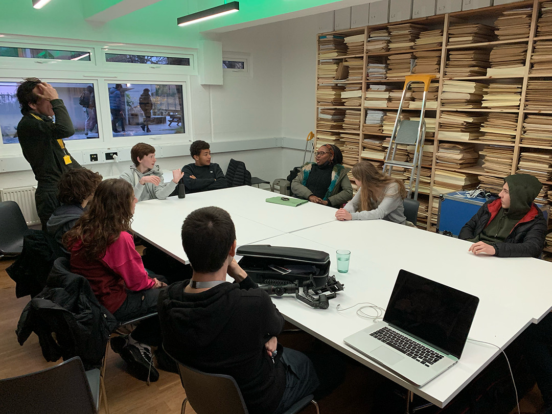 A group of people sits around a large white table in a brightly lit room. The table has a laptop and a camera on it. One person is standing, while others are seated, engaged in conversation. Stacks of papers or files are organised on shelves against the wall.