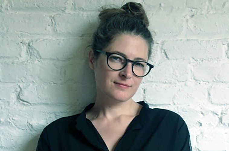 This is a headshot of Christine Sticher against a white brick wall. She wears a black shirt, glasses, her hair tied in a bun, and a slight smile.