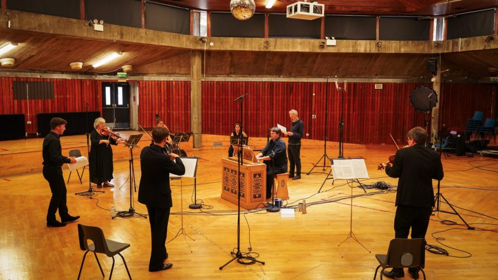 Orchestra players perform in a large, wood-panelled concert hall. A harpsichord player is seated in the centre, surrounded by violinists and a vocalist, with music stands and microphones set up around them.