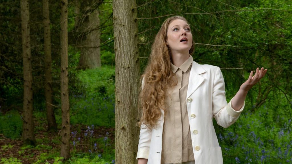 A woman with long curly blonde hair, wearing a white coat, beige shirt, and white pants, stands in a lush green forest. She looks up with one hand raised as if feeling for rain, surrounded by trees and vegetation.