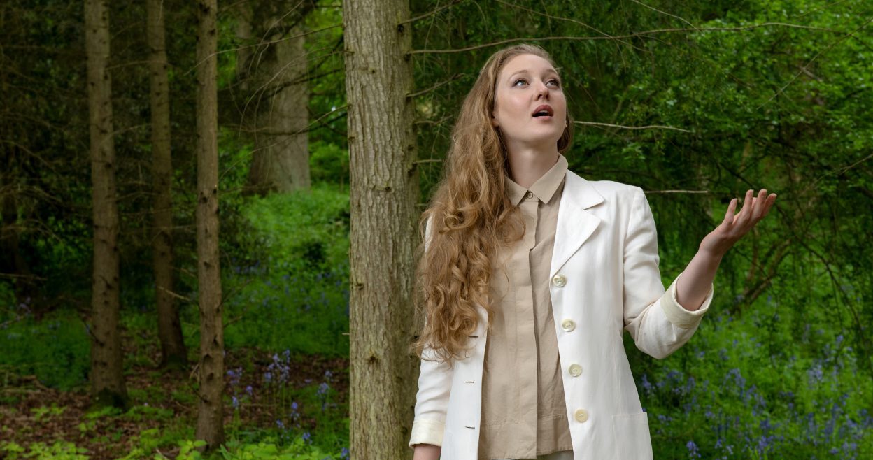 A woman with long curly blonde hair, wearing a white coat, beige shirt, and white pants, stands in a lush green forest. She looks up with one hand raised as if feeling for rain, surrounded by trees and vegetation.