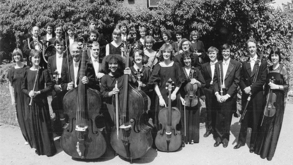 A black-and-white photo featuring a group of musicians posing outdoors. The ensemble includes string instrument players holding violins, cellos, and double basses. The backdrop is a lush green area with trees and a building partially visible. All individuals are smiling.