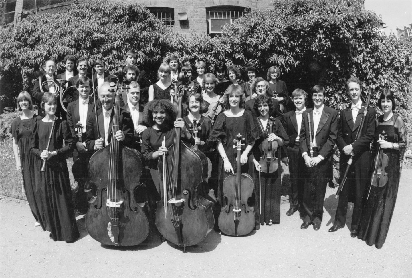 A black-and-white photo featuring a group of musicians posing outdoors. The ensemble includes string instrument players holding violins, cellos, and double basses. The backdrop is a lush green area with trees and a building partially visible. All individuals are smiling.