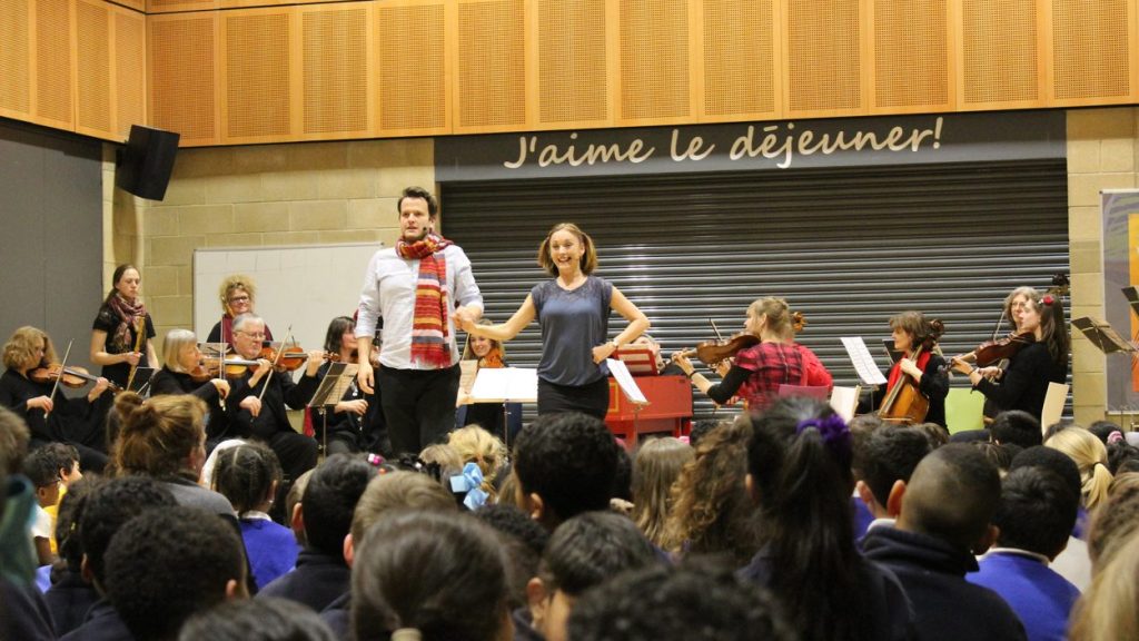 A man and a woman stand at the front of a classroom, engaging an audience of students. Behind them, a group of musicians play various string instruments. The wall displays the phrase "J'aime le déjeuner!" There is a vibrant, educational atmosphere.