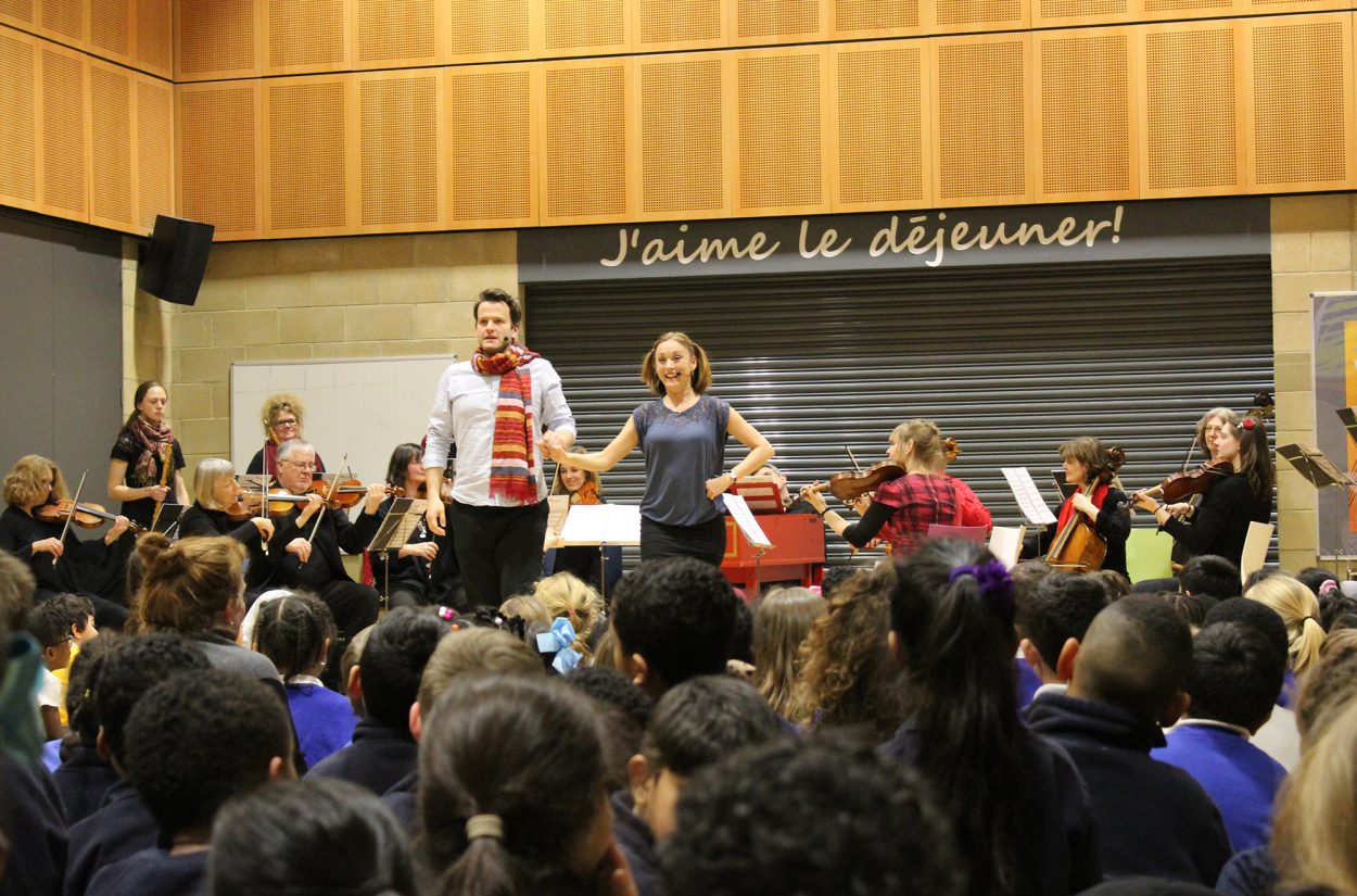 A man and a woman stand at the front of a classroom, engaging an audience of students. Behind them, a group of musicians play various string instruments. The wall displays the phrase "J'aime le déjeuner!" There is a vibrant, educational atmosphere.