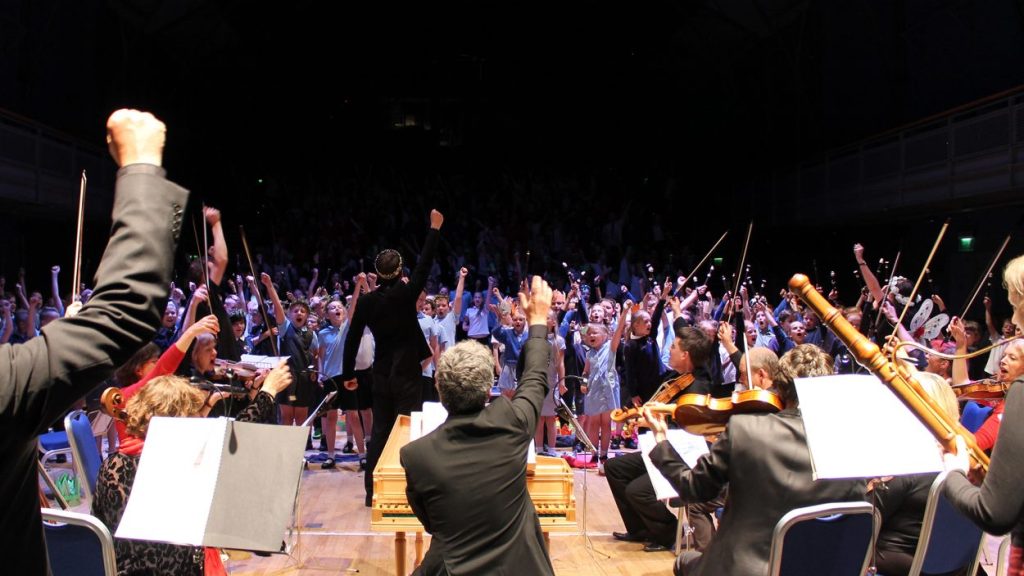 A symphony orchestra is performing on stage with the conductor in the centre. Musicians are playing various instruments, including violins and woodwinds. The audience in the background enthusiastically raises their hands in applause and cheer.