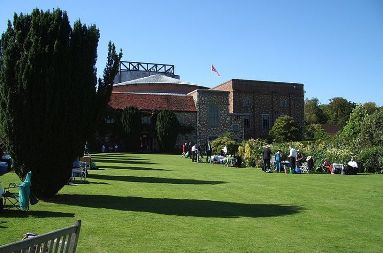 A brick building with a red roof sits at the end of a large, well-maintained lawn. Tall trees cast long shadows across the grass. Several people are enjoying the sunny day, sitting and standing in the shaded areas and along the perimeter of the building.