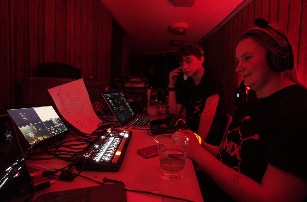 Two students operating video and sound equipment in a dimly lit room with red lighting. One student is sitting, wearing headphones, and holding a glass, while the other, also wearing headphones, is looking at multiple screens and control panels on a desk.