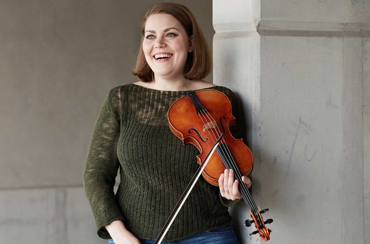 A smiling woman with shoulder-length brown hair is leaning against a white wall. She is holding a violin and bow. She is wearing a green knit sweater and jeans. The background is plain and neutral.