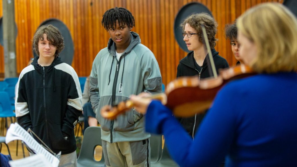 Four students stand in a music classroom with wooden-paneled walls, watching a Julia Kuhn play the violin. Empty chairs and music stands are visible in the background.