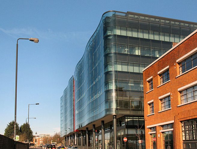 A modern glass-fronted building with a wavy design stands by the roadside. It is situated next to a traditional red-brick building. Both structures are under a clear blue sky, with minimal street elements including a lamppost and some trees visible in the distance.