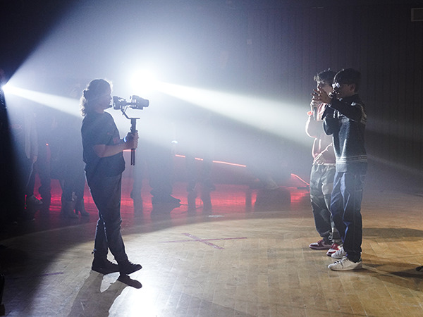 A videographer holds a camera stabiliser while filming two students on stage in a dimly lit room with beams of light shining through the haze. Onlookers stand in the background watching the scene unfold.