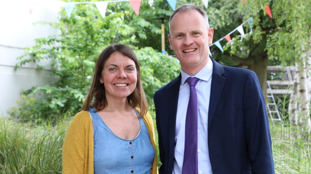 Nicholas John and Martha Collins stand in a garden with lush green vegetation and colourful bunting hanging. Nicholas wears a navy suit and purple tie, with Martha on the left wearing a yellow cardigan. They are both smiling.