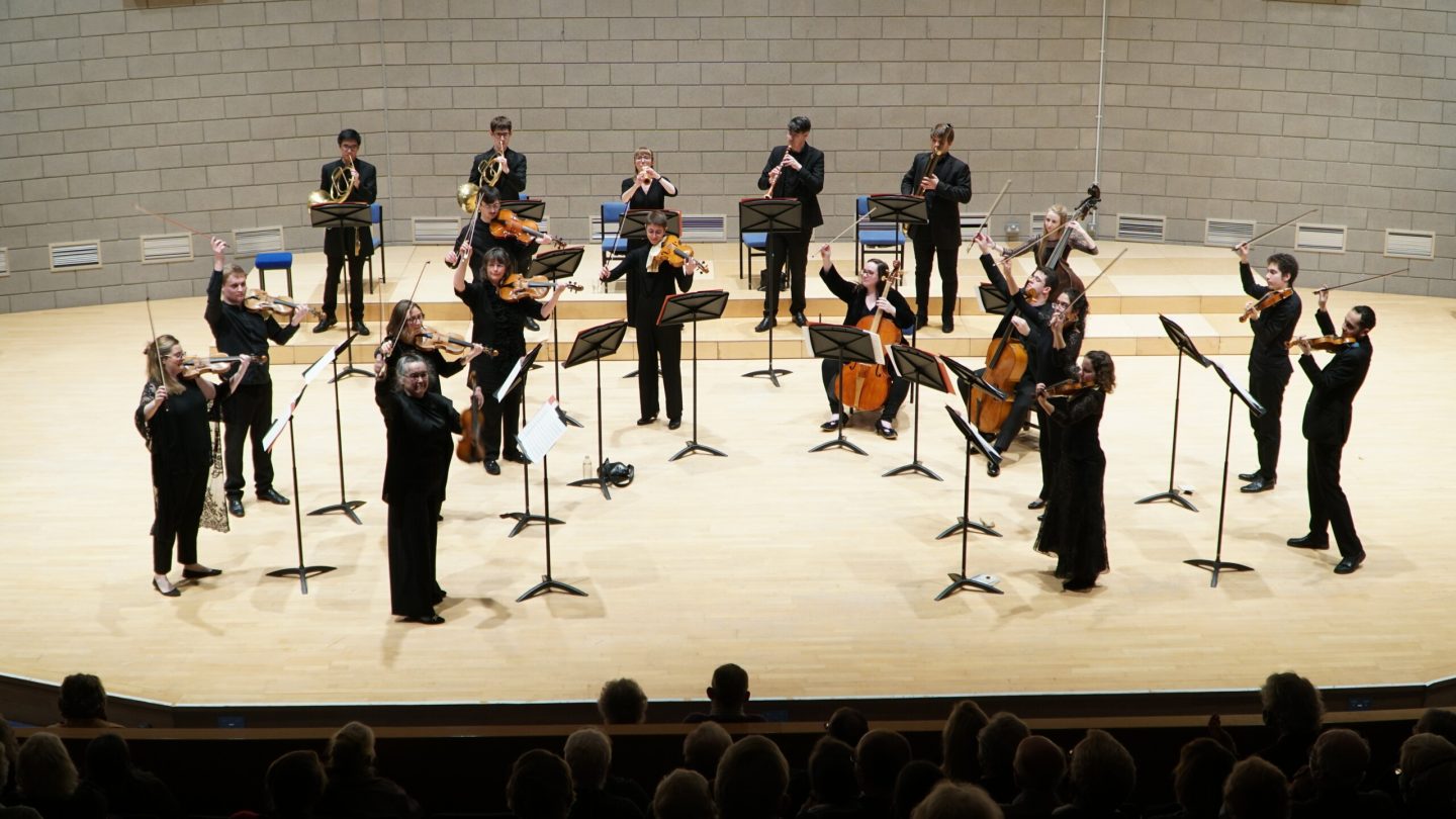 A group of musicians dressed in black perform in an orchestra on a stage. They are playing various instruments, including violins, cellos, and wind instruments. The conductor stands in front, guiding the performance. Audience members are visible in the foreground.