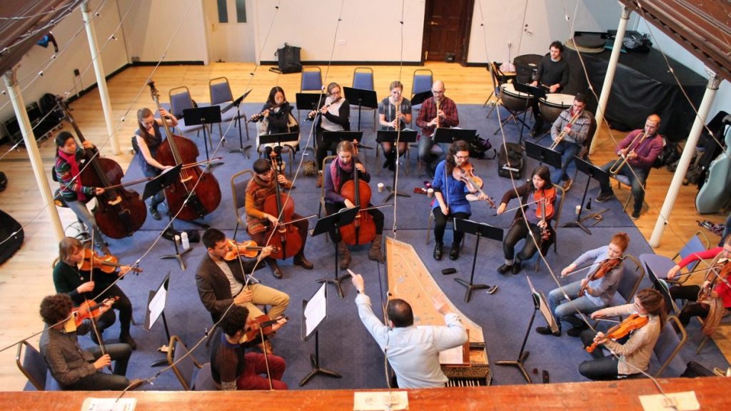 A group of musicians are in a rehearsal space playing various string and wind instruments, arranged in a semicircle around a conductor. The setting is a spacious room with wooden floors and music stands. The conductor stands at the centre facing the musicians.