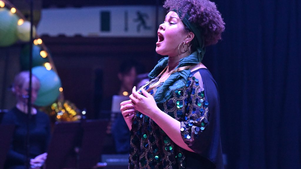 A woman with curly hair wearing a sparkly, sequinned outfit performs passionately on stage. She stands with her mouth open, singing. In the background, there are musicians and blurred stage decorations with green and yellow lights.