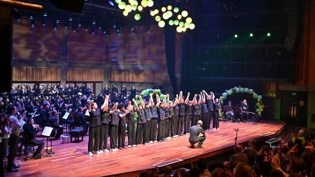 A group of performers in matching attire stand on stage holding hands and taking a bow, while an audience applauds. Behind them, an orchestra and more performers can be seen. The stage is lit with bright lights, and a cluster of balloons hangs overhead.