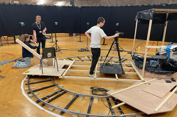 Several individuals work together on a circular track setup inside a large indoor space. One person adjusts a camera on a tripod, while another handles equipment nearby. The track, made from wooden panels and metal, is set on a gymnasium floor with scattered tools.