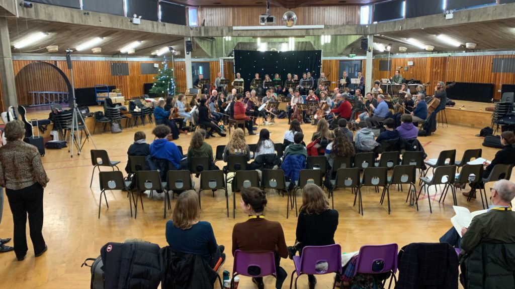 A group of musicians, both sitting in chairs and standing, rehearses in a large, open room with wooden walls and a high ceiling. Several people are seated in a semi-circle watching the rehearsal, and some in the audience take notes.