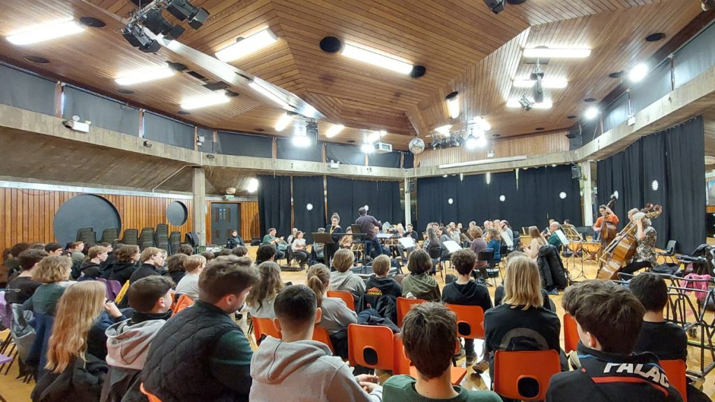 An audience of students and community members sit in orange chairs watching the orchestra players rehearse in a wood-panelled, brutalist hall with a high, vaulted ceiling and bright lights.