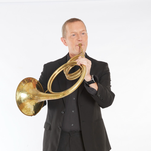 Roger Montgomery wears a black suit and stands in front of a white background. He holds a horn to his mouth and looks off camera as he plays.