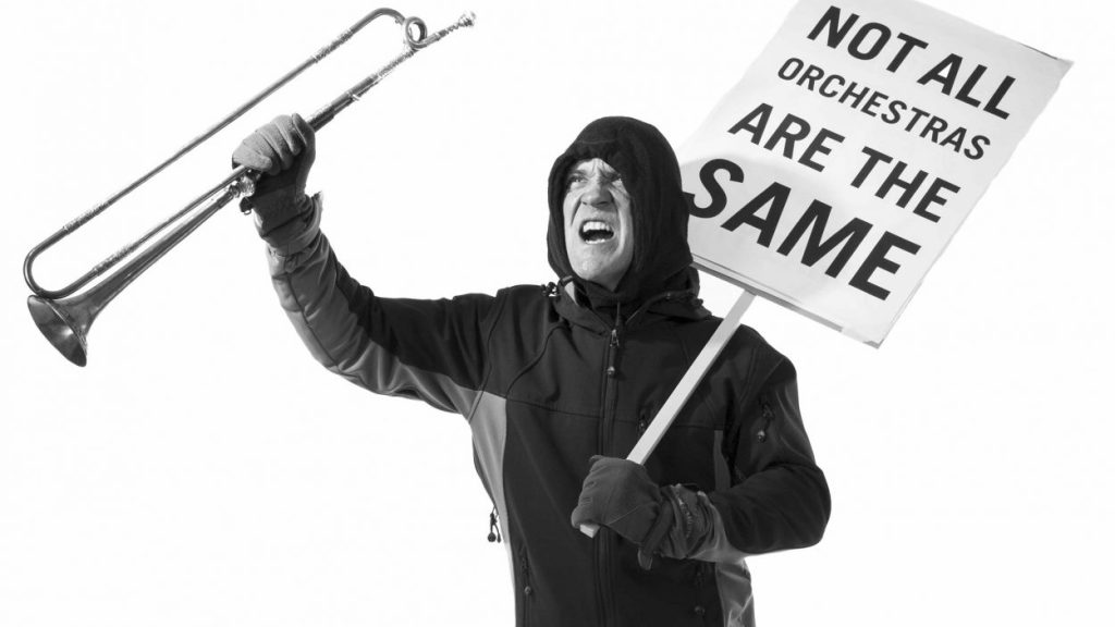 A person in a hooded jacket energetically holds up a sign that reads "Not all orchestras are the same" in one hand and a trumpet in the other. The image is in black and white, creating a dramatic and impactful scene.