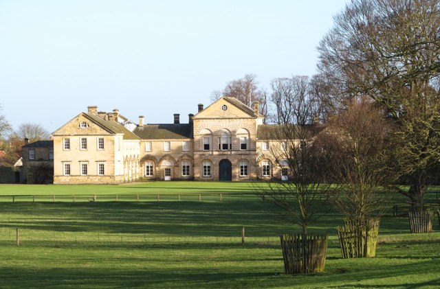 A large, historic mansion with a symmetrical facade, arched windows, and beige brickwork stands amidst a well-maintained lawn. Bare trees are scattered across the grassy foreground, and the entire scene is bathed in soft daylight.