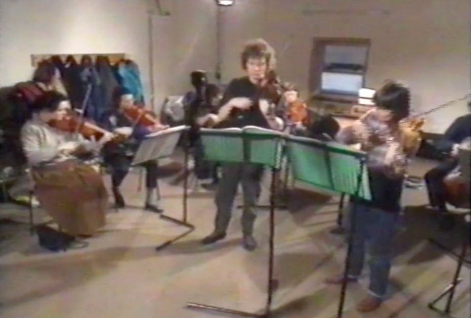 A group of people playing various string instruments in a rehearsal room. Music stands with green sheet music are in front of the musicians. Coats hang on hooks along the back wall. The room has beige walls and a rectangular window.