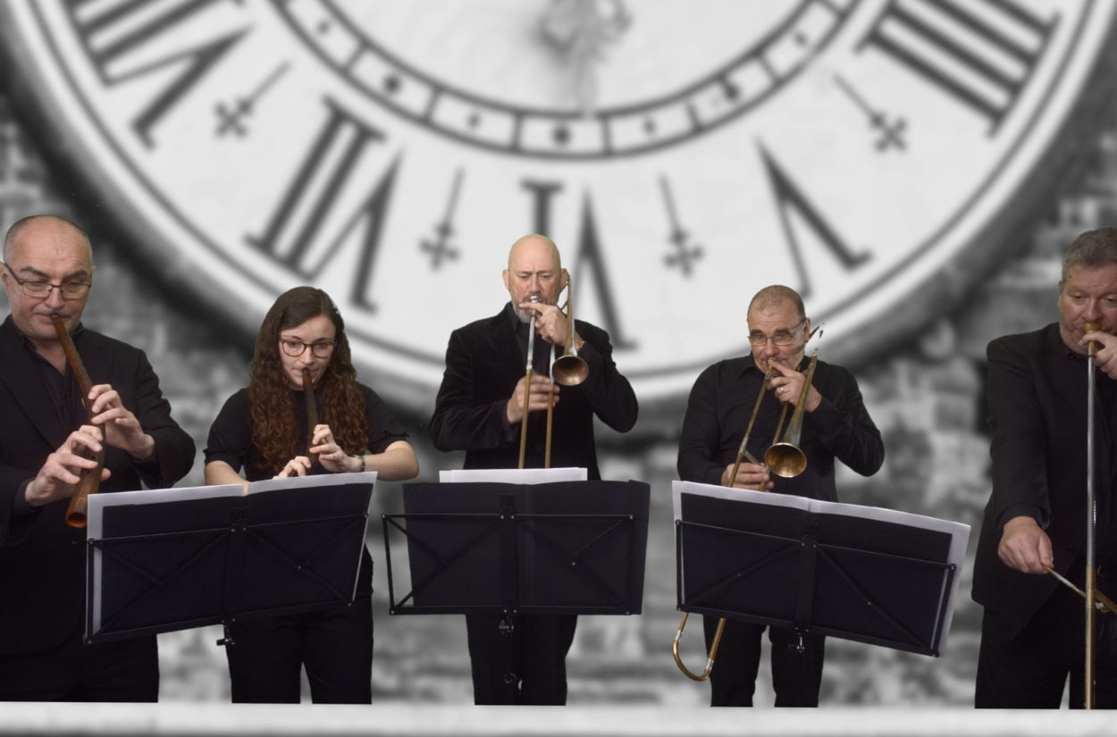 Five musicians play brass instruments in front of music stands, standing against a backdrop featuring a large, black-and-white clock face with Roman numerals. They are dressed in black attire and are focused on their performances.