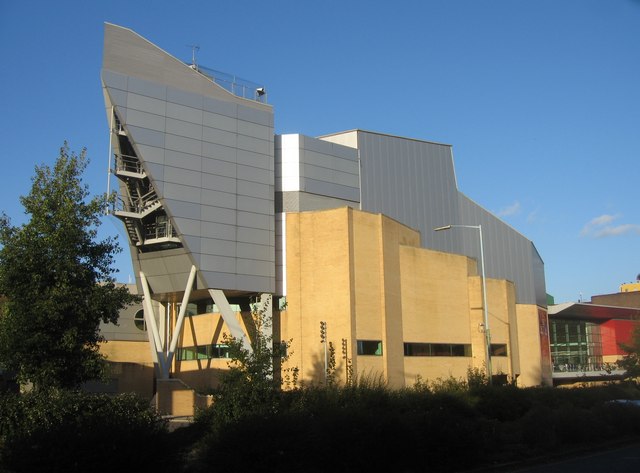 Modern building with an angular, metallic design on the upper level and a brick base, surrounded by shrubs and trees. The structure features large, geometric shapes and a combination of glass and metal panels. The sky is clear and blue.