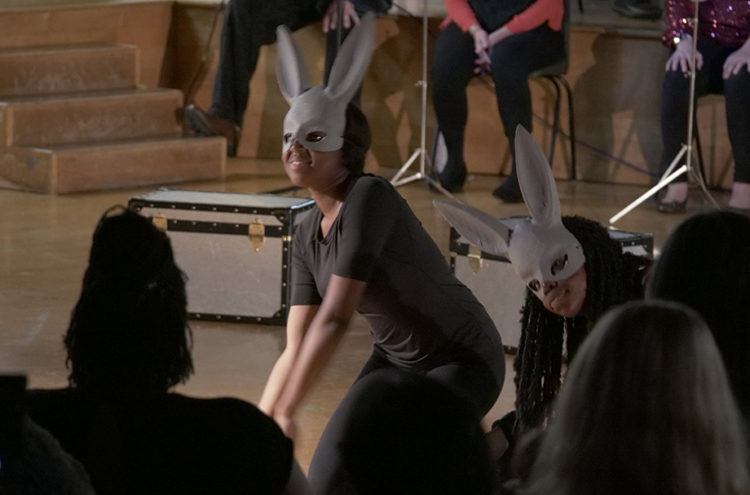 Two performers wearing rabbit masks and black outfits, crouch on a wooden floor, mimicking rabbit movements during a theatre performance. Seated audience members watch in dim lighting. A large black and white trunk is visible in the background near wooden steps.