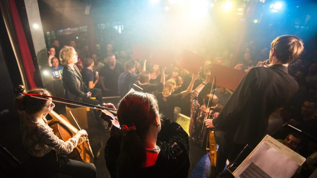 A musical performance is taking place on a dimly lit stage. Musicians, including a cellist and violinist, are playing while a singer performs. The audience is seen in the background, standing and attentively watching the show, with colourful lights illuminating the scene.