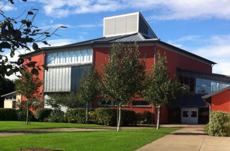 A modern, red-painted building with large windows sits amidst a landscaped area with green lawns and young trees. The sky above is partly cloudy, and a paved walkway leads to the entrance. The building's design features a mix of flat and angled roofs.