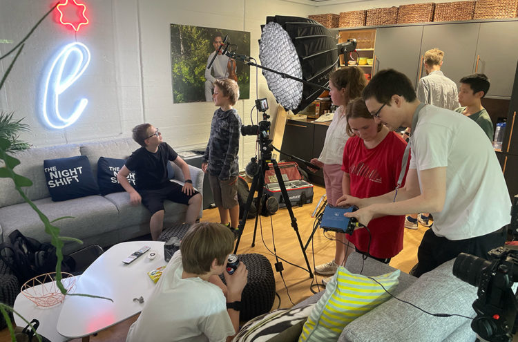 A group of students are on a film set. Several individuals are setting up cameras and lighting equipment, while others are sitting and talking. The room features a sofa with "The Night Shift" pillows, leafy plants, and a neon light on the wall.